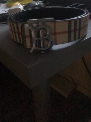  Burberry bälte  - Snyggt bälte från Burberry med klassiskt rutigt mönster i beige, svart och rött. Bältet har ett stort metallspänne med ett B framtill. Perfekt accessoar för att lyfta din outfit. Kan va både svart och vanlig.