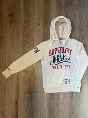 Superdry Athletics grå hoodie - Ljusgrå hoodie från Superdry med trycket 'Superdry Athletics Track. JPN' i rött och svart på bröstet🤗 Passar Xs🙌🏻 pris ej hugget i sten😇
