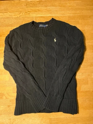 Svart kabelstickad tröja Polo Ralph Lauren - Kabelstickad svart tröja från Polo Ralph Lauren med rund halsringning och ribbade muddar. Klassisk broderad logga på bröstet i vitt. Perfekt för lager på lager och enkel att matcha med jeans eller kjol.