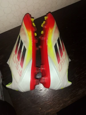 Adidas F50 vita fotbollsskor - Säljer ett par Adidas F50 fotbollsskor i vitt med röda, svarta och gula detaljer. Skorna har en strömlinjeformad design, låg vikt och mönstrad ovandel för bättre bollkontroll. Yttersulan är röd med fasta dobbar, perfekt för spel på gräsplan. Snygg och modern look för dig som vill sticka ut på planen.