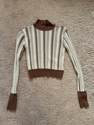 Randig ribbad polotröja brun/beige - Säljer en ribbad långärmad tröja med polokrage i beige och brunt. Tröjan har vertikala ränder, breda muddar vid ärmslut och midja samt en croppad passform. Perfekt för dig som gillar retrovibbar och stilrena färger.