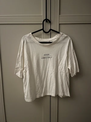 Vit t-shirt med texttryck - Vit t-shirt i bomull med svart texttryck 'GOOD VIBES ONLY' på bröstet. Klassisk rund halsringning och korta ärmar. Perfekt för en avslappnad och enkel stil.