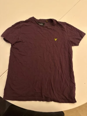 Lila t-shirt från Lyle & Scott - Snygg lila t-shirt från Lyle & Scott med klassisk gul logga broderad på bröstet. T-shirten har rund hals och korta ärmar, perfekt för en avslappnad stil. Tillverkad i mjukt bomullsmaterial som känns skönt mot huden.