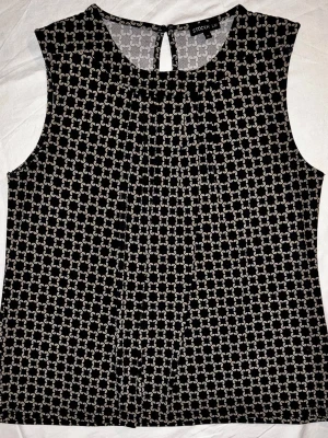 Mönstrad blus från Stockh lm (MQ) - Klassisk och stilren ärmlös blus från märket Stockh lm (säljs hos MQ Marqet). Toppen har ett fint geometriskt mönster i svart och vitt och dekorativa veckdetaljer framtill som ger ett snyggt fall. Perfekt att bära under en kavaj på kontoret eller till ett par snygga byxor för en mer klädd vardagslook. Detaljer: Rund halsringning, slits med knapp i nacken. Mått: Bredd: ca 50 cm Längd: ca 61 cm.