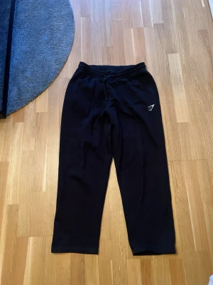 Svarta mjukisbyxor från Gymshark - Svarta mjukisbyxor från Gymshark med avslappnad passform och dragsko i midjan. Byxorna har en diskret logga på ena benet och är tillverkade i mjukt material som passar perfekt för chill eller träning.