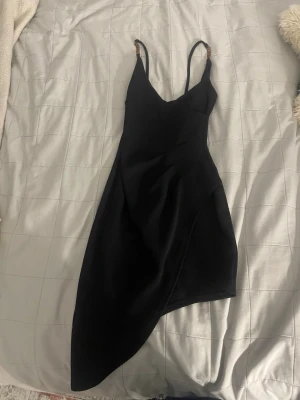 Svart asymmetrisk klänning NLY One - Säljer en svart kortklänning från NLY One i storlek XS. Klänningen har smala axelband med gulddetaljer och en snygg asymmetrisk nederkant som ger en unik look. Perfekt för dig som vill sticka ut lite extra.