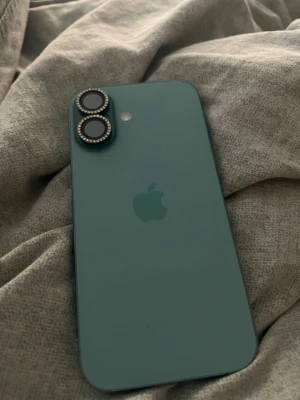 Blå iPhone 16 - Snygg blå iPhone med två kameror på baksidan och stilren matt finish. Telefonen har Apples klassiska logga och platt design med tydliga knappar på sidan. Kameraringarna är dekorerade med små stenar för extra bling.