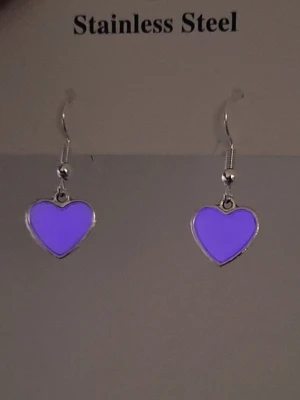 Glow in the dark hjärtformade örhängen Vit/Lila - Säljer ett par söta örhängen i rostfritt stål med hängande hjärtan som är vita i dagsljus och lyser lila med ett tydligt sken i mörker efter att ha varit en kort stund i vanligt dagsljus eller solljus. Hjärtana har en cool glow-effekt i mörker som varar i flera timmar efter normal användning och örhängena har klassisk krok i rostfritt stål. Perfekta för dig som vill sticka ut med färg och form! Tål vatten & har en mycket lång livslängd. Handgjorda.