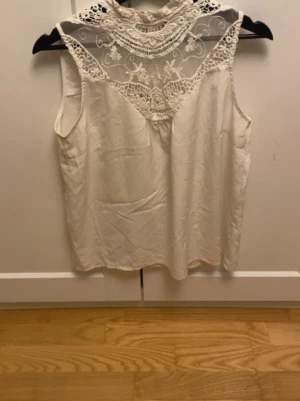 Vit spetsdetaljerad ärmlös topp - Snygg vit ärmlös topp med vackra spetsdetaljer och broderier upptill. Toppen har hög hals och transparent spets över axlar och bröst, vilket ger en romantisk och elegant look. Perfekt att styla med jeans eller kjol.