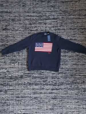 Mörkblå stickad tröja Ralph Lauren - Mörkblå stickad tröja från Ralph Lauren med amerikansk flagga i rött, vitt och blått på bröstet samt RL-broderi. Lappen är inte fast men kan skicka den med!