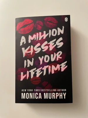A Million Kisses In Your Lifetime - En romantisk roman av Monica Murphy med svart omslag dekorerat med röda och rosa läppavtryck. Boken är en pocketutgåva och har tydlig, vit text på framsidan. Perfekt för dig som gillar kärlekshistorier och ungdomsromaner. Helt ny, har bara stått på bokhyllan