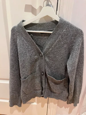 Grå stickad kofta med fickor - Mysig grå stickad kofta med v-ringning och tre knappar framtill. Koftan har två stora fickor och ribbade muddar vid ärmslut och nederkant. Perfekt för lager på lager och enkel att matcha med olika outfits.