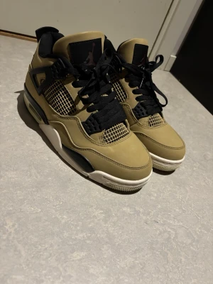 Air Jordan 4 sneakers beige/svart - Säljer ett par Air Jordan 4 sneakers i beige mocka med svarta detaljer och meshpaneler. Skorna har svart snörning, vit och svart sula samt ikonisk Jumpman-logga på plösen. Perfekta för dig som vill sticka ut med en klassisk och stilren look.