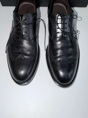 Svarta loafers i skinn med broguemönster - Säljer ett par svarta loafers i äkta skinn med klassiskt broguemönster och snörning. Skorna har en elegant, spetsig tå och dekorativa perforeringar längs kanterna. Perfekta för dig som vill ha en stilren och tidlös look.