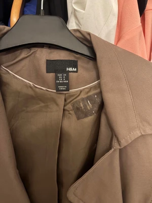 Brun trenchcoat från H&M - Stilren brun trenchcoat från H&M med klassisk krage, knytskärp i midjan och dekorativa knappar. Perfekt för dig som vill ha en tidlös och trendig look.