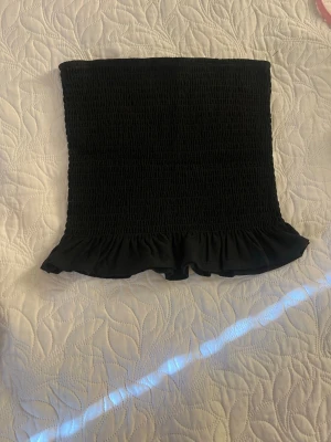 Svart smockad tubtopp med volang - Säljer en svart tubtopp med smockad resår och söt volangkant nedtill. Toppen är axelbandslös och har en tight passform som sitter snyggt på kroppen. Perfekt att styla till jeans eller kjol för en trendig look.