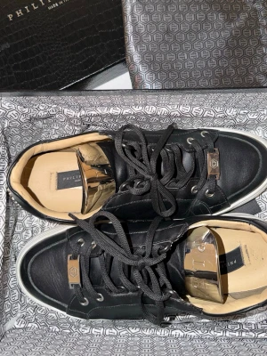 Svarta Philipp Plein sneakers i läder - Snygga svarta sneakers från Philipp Plein i äkta läder med vita sulor och coola metalldetaljer på snörningen. Insidan är beige och skon har en exklusiv känsla med logotyp på plösen och hälen. Perfekta för dig som gillar lyxiga och trendiga skor. All tillbehör följer med!