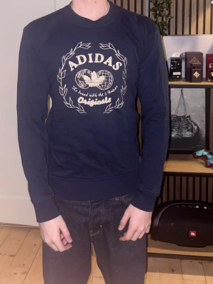 Mörkblå sweatshirt från Adidas - Mörkblå sweatshirt från Adidas Originals med tryck framtill i vitt och beige. Storleken är XS men den passar mest S skulle jag säga. Tröjan har rund halsringning, långa ärmar och ribbade muddar vid ärmslut och nederkant. Tillverkad i mjukt bomullsmaterial för en bekväm känsla. 