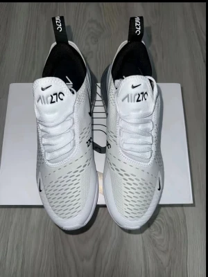 Nike Air 270 vita sneakers - Fräscha vita Nike Air 270 sneakers med svart logga och detaljer. Ovandelen är i meshmaterial för bra andningsförmåga och komfort. Snygg design med dragflik bak och diskreta mönster på sidorna. Perfekta för en sportig och trendig look.