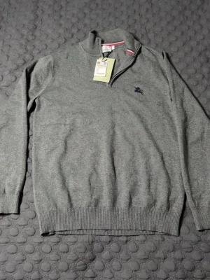 Grå halvzip tröja från Burberry - Snygg grå stickad tröja från Burberry med halv dragkedja och ribbade muddar. Tröjan har en diskret broderad logga på bröstet och en randig detalj på insidan av kragen. Perfekt för dig som gillar klassisk och stilren design. STORLEK M, PASSAR S. HELT NY ALDIRG ANVÄND. 