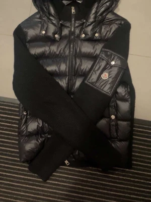 Moncler Cardigan ÄKTA - Snygg svart dunväst från Moncler med glansig finish, huva och stickade svarta ärmar. Den är storlek L men passar M!
