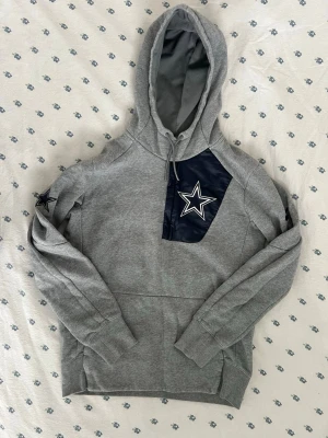 Grå Dallas Cowboys hoodie från Nike - Snygg grå hoodie från Nike med Dallas Cowboys stjärnlogga på bröstet och ärmen. Hoodien har en stor huva med dragsko, magficka och detaljer i mörkblått tyg. Tillverkad i mjukt sweatshirtmaterial, perfekt för dig som gillar sportig stil. Storlek M / medium. Importerad från usa. 