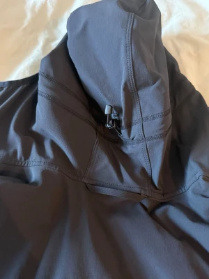 Svart vindjacka från Haglöfs med huva - Snygg svart vindjacka från Haglöfs med justerbar huva och meshfoder på insidan. Jackan har minimalistisk design, diskret logga på bröstet och är tillverkad i lätt, vindtätt material. Perfekt för blåsiga dagar och outdoor-aktiviteter.