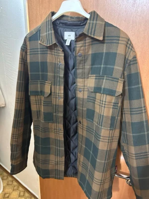 Rutig overshirt från H&M - Snygg overshirt från H&M i grönt och brunt rutigt mönster. Jackan har två bröstfickor med lock, tryckknappar framtill och en quiltad svart insida. Perfekt lager på lager-plagg med avslappnad passform och klassisk krage.