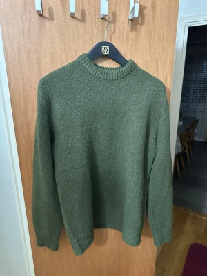 Olivgrön stickad tröja med rund hals - En enkel och stilren olivgrön stickad tröja med rund halsringning och ribbade muddar vid ärmslut och nederkant. Perfekt för lager-på-lager och passar till det mesta. Tröjan har en klassisk passform och är mjuk och skön.