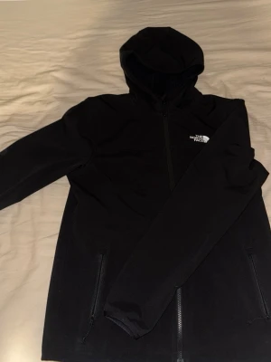 Svart vindjacka från The North Face - Snygg svart vindjacka från The North Face (använd 3-5 gånger) med huva och dragkedja framtill. Jackan har två fickor med dragkedja och logga på bröstet samt ryggen. Perfekt för dig som vill ha en stilren och funktionell jacka som är vatten och vind  avvisande 