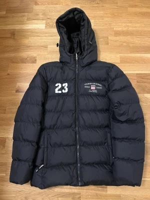 Svart pufferjacka från Vinson - Snygg svart pufferjacka från Vinson med huva och dragkedja framtill. Jackan har tryckta detaljer med siffran 23 och Vinson Original-logga på bröstet. Tillverkad i slitstarkt syntetmaterial och fodrad för extra värme. Perfekt för kalla vinterdagar. Pris kan diskuteras. Den har änvänds 3 gånger.