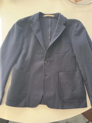 Marinblå kavaj från Dondup - Stilren mörkblå kavaj från Dondup med klassisk krage, tre utanpåliggande fickor och tvåknäppning. Tillverkad i ett mjukt tyg som ger en avslappnad men ändå dressad look. Perfekt för dig som vill ha en modern och enkel kavaj med snygga detaljer. Passar M-L.