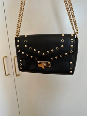 Svart axelväska Michael Kors nitar - Snygg svart axelväska från Michael Kors i slätt läder med guldfärgade nitar och kedja. Väskan har ett coolt mönster av runda nitar på locket och ett guldfärgat vridlås framtill. Perfekt för dig som gillar en edgy och trendig stil.