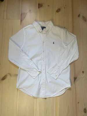 Ralph Lauren skjorta  - Klassisk vit skjorta från Ralph Lauren med broderad logga på bröstet. Skjortan har lång ärm, knappar framtill och vid manschetterna samt en tidlös krage. Perfekt till både jeans och chinos för en clean look. Jag är 169 cm på bild. Hör av er vid minsta fundering👍