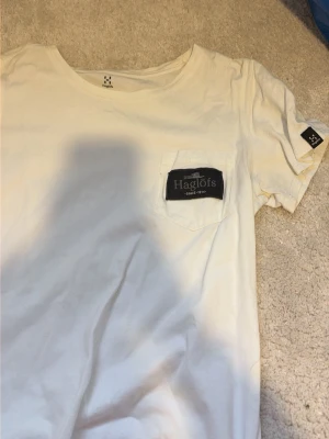 Vit t-shirt från Haglöfs med ficka - Vit t-shirt från Haglöfs med rund halsringning och en bröstficka med svart Haglöfs-logga. Klassisk och enkel design, perfekt till vardags. Tillverkad i mjuk bomull och har korta ärmar samt diskret logga även på ärmen.