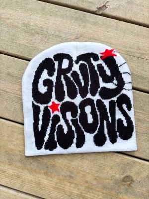 Vit mössa med GRvty VISIONS-tryck - Säljer en vit mössa med svart text 'GRvty VISIONS' i stor, grafisk stil och två röda stjärnor som detaljer. Kolla mina andra annonser för samfrakt. Mössan är stickad och har en avslappnad passform, perfekt för dig som gillar streetwear och vill sticka ut.