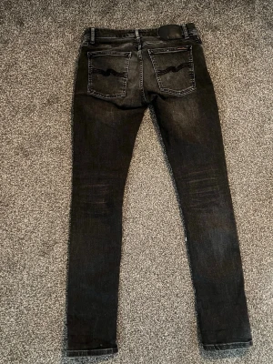 Svarta nudie jeans i modell skinny lin - Säljer ett par svarta skinny jeans med dragkedjegylf och klassisk femficksdesign. Jeansen har en tight passform och är tillverkade i ett stretchigt material som sitter snyggt. Perfekta för dig som gillar en slimmad look. Nudie jeans Skinny Lin