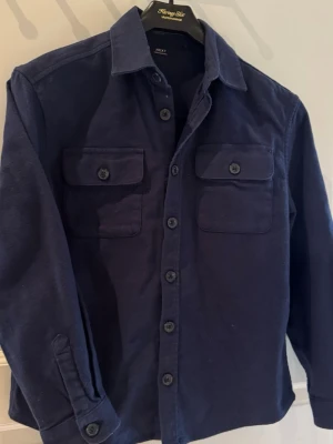 Mörkblå overshirt från Next - Stilren mörkblå overshirt från Next med raka linjer och klassisk krage. Två bröstfickor med lock och knapp, samt knäppning hela vägen framtill. Tillverkad i ett mjukt, lite tjockare tyg som ger en avslappnad men snygg look. Perfekt att bära öppen eller stängd.