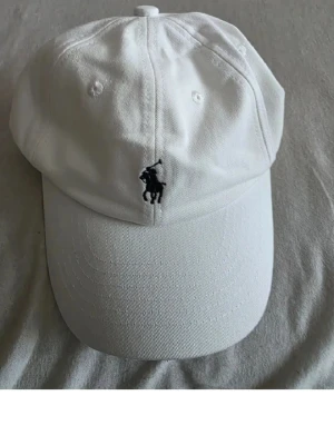 Vit keps från Polo Ralph Lauren - Snygg vit keps från Polo Ralph Lauren med klassisk böjd skärm och broderad svart logga med polospelare framtill. Justerbar rem baktill för perfekt passform. Tillverkad i bomull för en skön och luftig känsla.