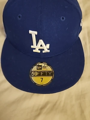 Blå LA Dodgers keps från New Era - Säljer en blå 59FIFTY keps från New Era med LA Dodgers-logga broderad i vitt framtill. Klassisk flat brim och broderad New Era-logga på sidan. Kepsen är i fast modell och har ventilationhål upptill. Perfekt för dig som gillar sportig streetstyle.