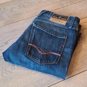 Replay Anbass - Snygga Replay Anbass jeans i klassisk blå tvätt med raka ben och lite mer slim passform. Jeansen har fem fickor, coola röd/rosa sömmar på bakfickorna och detaljerad Replay Blue Jeans-label i midjan. Perfekt för dig som gillar stilrena och tidlösa jeans. Jeansen är i nyskick då de knappt användts. De är L32 W32, det är bara att skicka om frågor