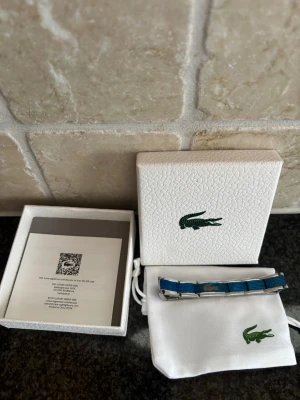 Blått lacoste armband - Säljer ett lacoste armband i nyskick!🐊 Blått med silvriga kanter och insida. Box och allt ingår. Ca 20 cm (justerbart). Repfri och rostfri. 