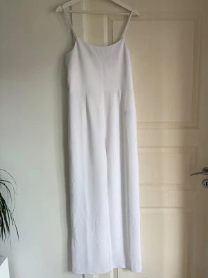 Vit ärmlös jumpsuit med raka ben - Säljer en stilren vit jumpsuit med smala axelband och raka, vida ben. Jumpsuiten har en enkel och clean design som ger en modern look. Perfekt för dig som gillar minimalistisk stil och vill ha något bekvämt men ändå snyggt.