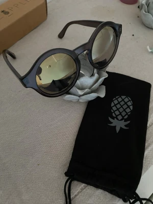 Runda svarta solglasögon Pineapple - Snygga runda solglasögon från Pineapple med svart båge och spegelglas. Kommer med en svart tygpåse med ananastryck. Perfekta för soliga dagar och ger en cool retrovibe till din stil.