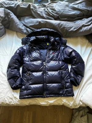 Mörkblå dunjacka från Polo Ralph Lauren - Snygg mörkblå dunjacka från Polo Ralph Lauren med blank finish och avtagbar huva. Jackan har två dragkedjeförsedda fickor, hög krage och diskret röd logga på bröstet. Perfekt för kalla vinterdagar och riktigt varm tack vare dunfyllningen. 