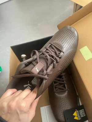 Bruna sneakers från Puma - Snygga bruna sneakers från Puma med perforerat mönster på ovandelen och klassisk snörning. Skorna har en sportig look med meshdetaljer på plösen och diskret Puma-logga. Perfekta för dig som gillar stilrena och bekväma sneakers.
