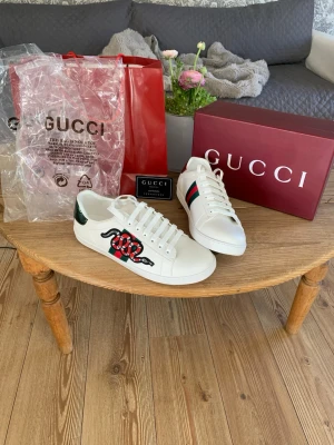 Gucci Ace sneakers - Vita Gucci Ace sneakers i läder med ikonisk grön och röd sidoremsa samt broderad röd orm på sidan. Metallicröd detalj på ena hälen och metallicgrön på den andra. Klassisk låg modell med vita snören och gummisula. Perfekt för dig som vill sticka ut.