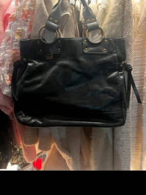 Svart handväska med gulddetaljer - Kenneth Cole Reaction handväska i äkta läder Snygg och rymlig svart handväska i mellanstorlek. Tillverkad i äkta läder, med guldfärgade detaljer och dubbla handtag. Gott skick, inga större märken eller skador. Perfekt vardagsväska som även rymmer mindre laptop/surfplatta. Nypris ca 1500–2000 kr.