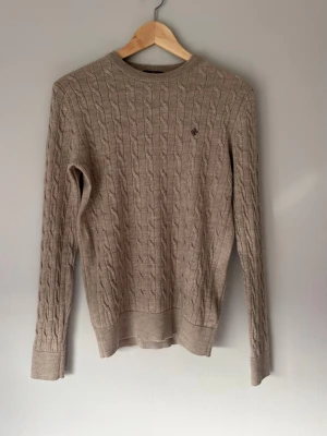 Beige kabelstickad Morris tröja  - Snygg beige kabelstickad tröja från Morris i 100% merinoull med rund halsringning och diskret logga broderad på bröstet. Tröjan har långa ärmar och ribbade muddar vid ärmslut och nederkant. Perfekt för lager på lager och klassisk stil.