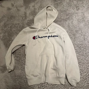 Beige hoodie från Champion - Säljer en beige hoodie från Champion med klassisk logga broderad i blått och rött på bröstet. Hoodien har stor huva med dragsko och en stor magficka framtill. Perfekt för en avslappnad och sportig stil.